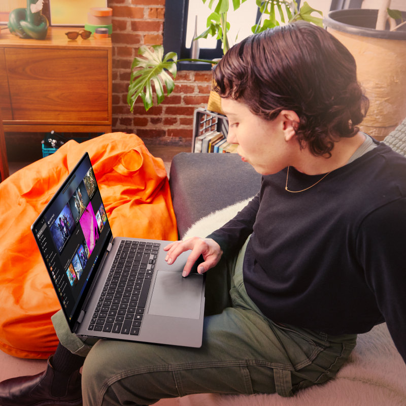 A person sitting on a sofa looking at PC games on an open laptop with a bean bag, amp, shelf and potted plant in the background.